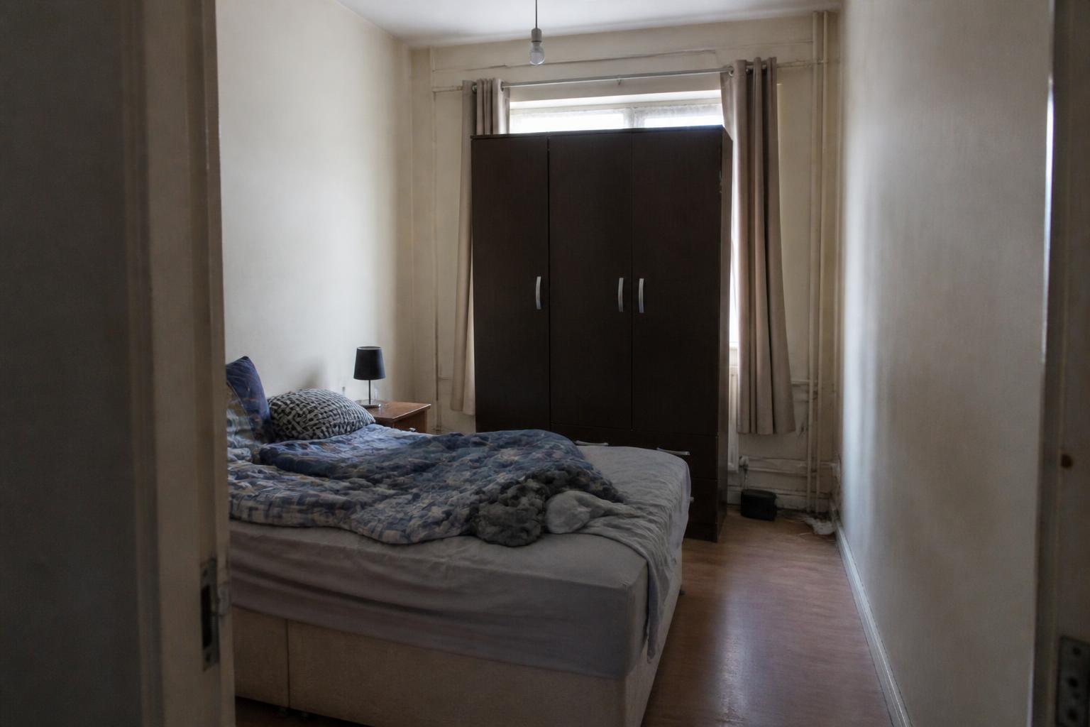Study of a cramped bedroom before: scuffed magnolia walls, bulky dark wardrobe blocking the window, worn laminate floor