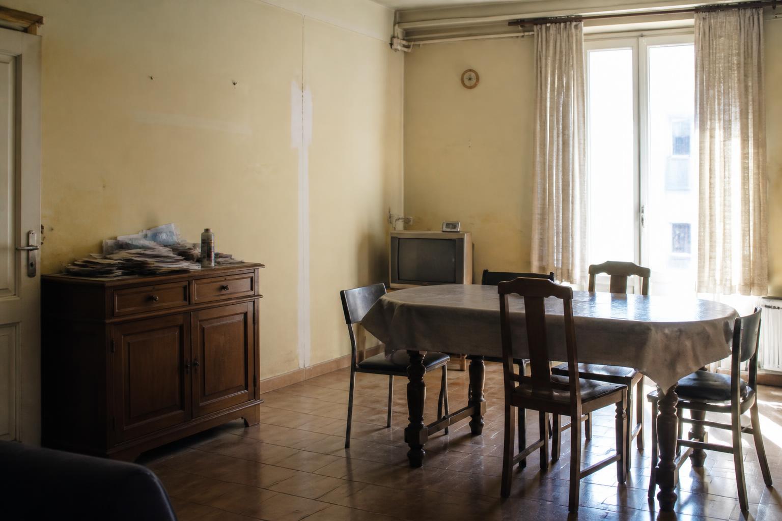 Study of a tired dining corner before renovation: yellowed cracked plaster, dark heavy table, plastic cloth and a tired curtain over the tall window