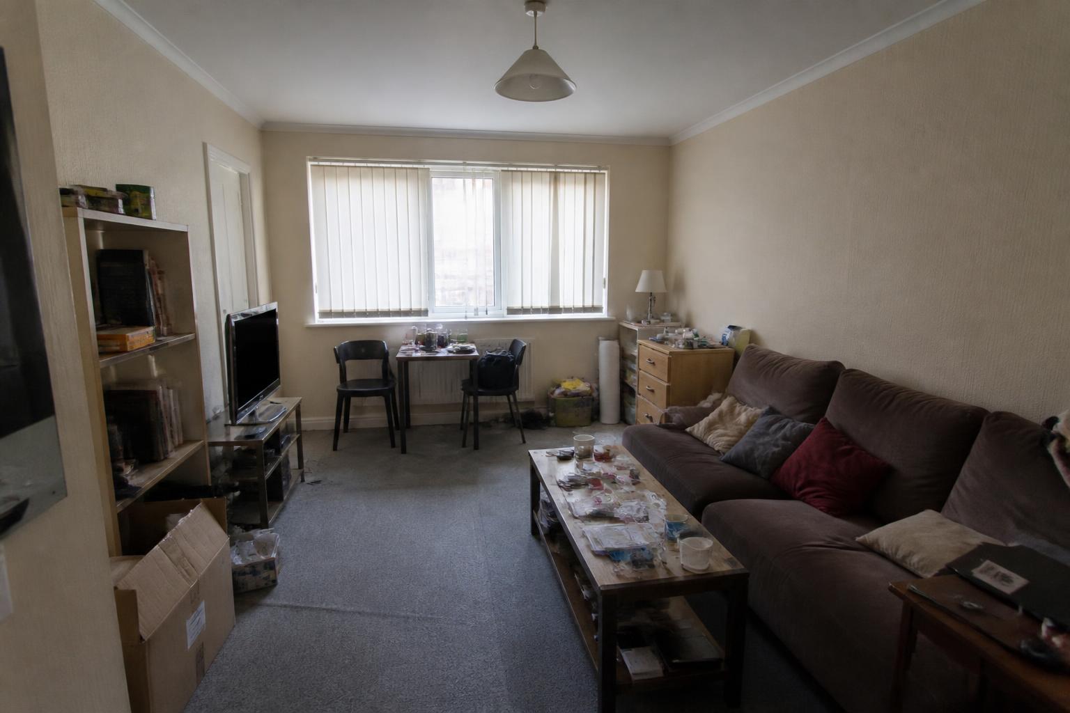 Study of a dated studio living room before: beige walls, worn carpet, oversized sofa and clutter under a single ceiling light